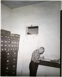 A man writing in a room with many boxes and filing cabinets