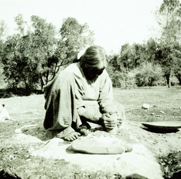 Indian woman grinding with pestle in a mortar, basketry tray beside her