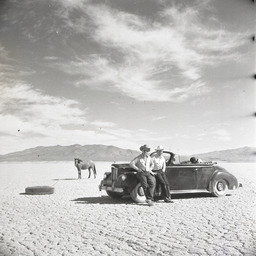 Men posing by car with horse roped to tire in background