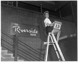 Diane Schindler on a ladder with a Friday the 13th calendar page in front of 'The Riverside Reno' sign, 2