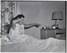 A woman in a nightgown in bed stretches for an alarm clock