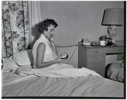 A woman in a nightgown uses a phone and holds up a clock while sitting in bed