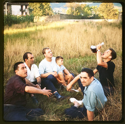 Group of men sitting in grass, one drinking from a glass wine pitcher