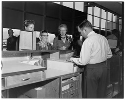 Three women at a counter with a man behind it assisting them, 1