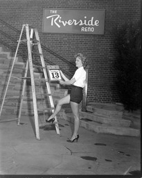 Diane Schindler on a ladder with a Friday the 13th calendar page in front of 'The Riverside Reno' sign, 1