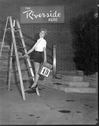 Diane Schindler on a ladder with a Friday the 13th calendar page in front of 'The Riverside Reno' sign, 3