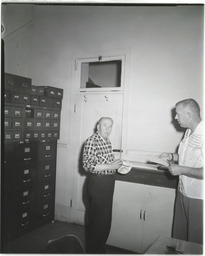 A man writing in a room with many boxes and filing cabinets with another man